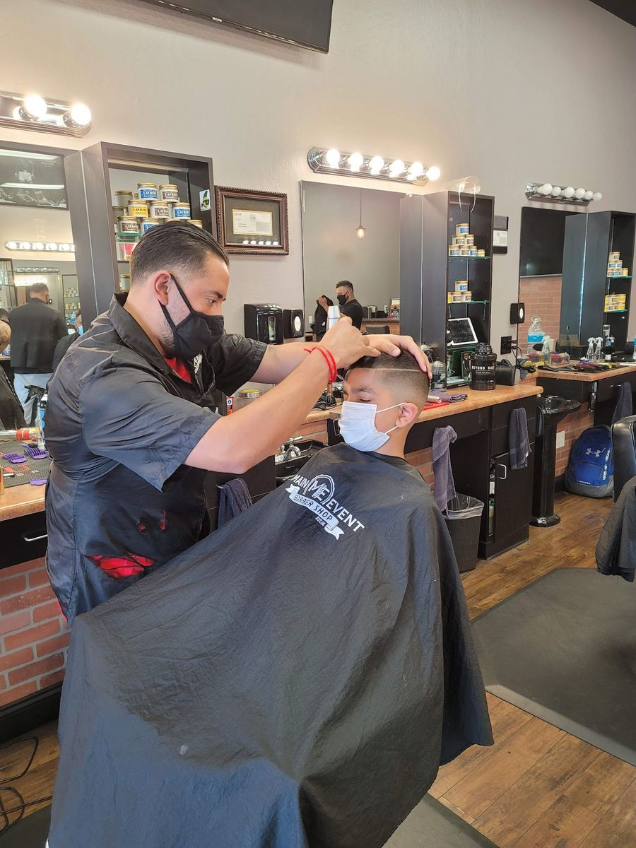 A barber in a black shirt and mask clipping a young client in a black Main Event cape, with mirrored stations behind