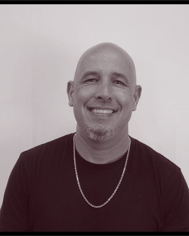 Portrait of Jeremy Carrier, owner of The Main Event Barber Shop, smiling, in a black T-shirt and a silver chain