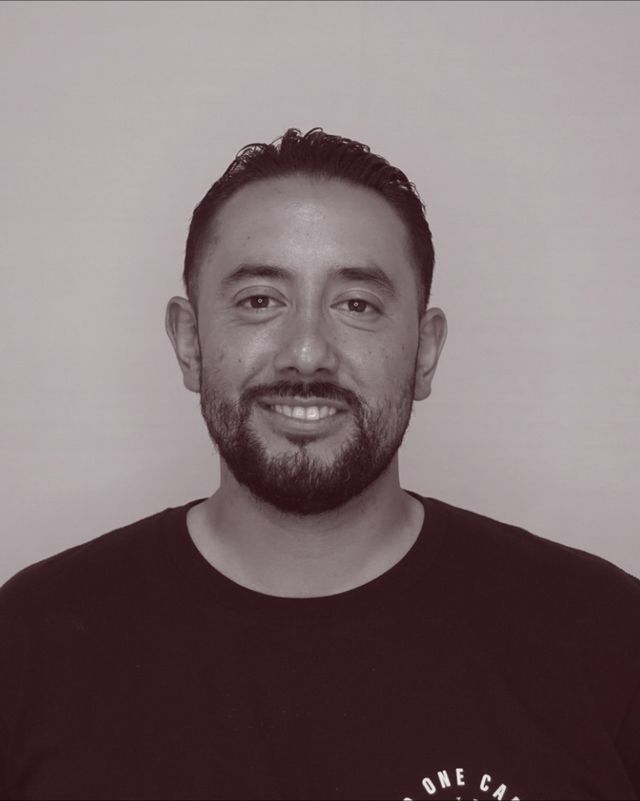 Portrait of barber Rolando “Fran” Zamora, smiling, with a full beard, in a black T-shirt