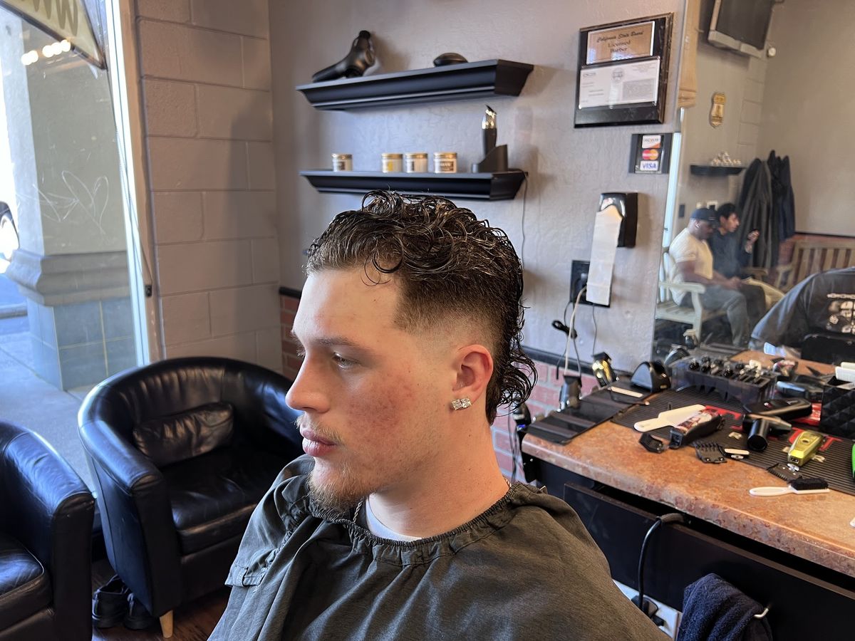 A young man in the chair with a curly top, a low fade and a lined beard, product shelves behind him