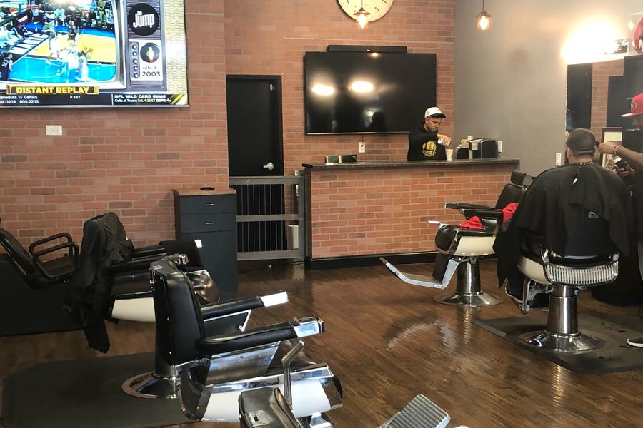 Inside The Main Event Barber Shop: black and chrome barber chairs on a wood floor, red brick walls, a wall clock, TVs and a brick bar counter