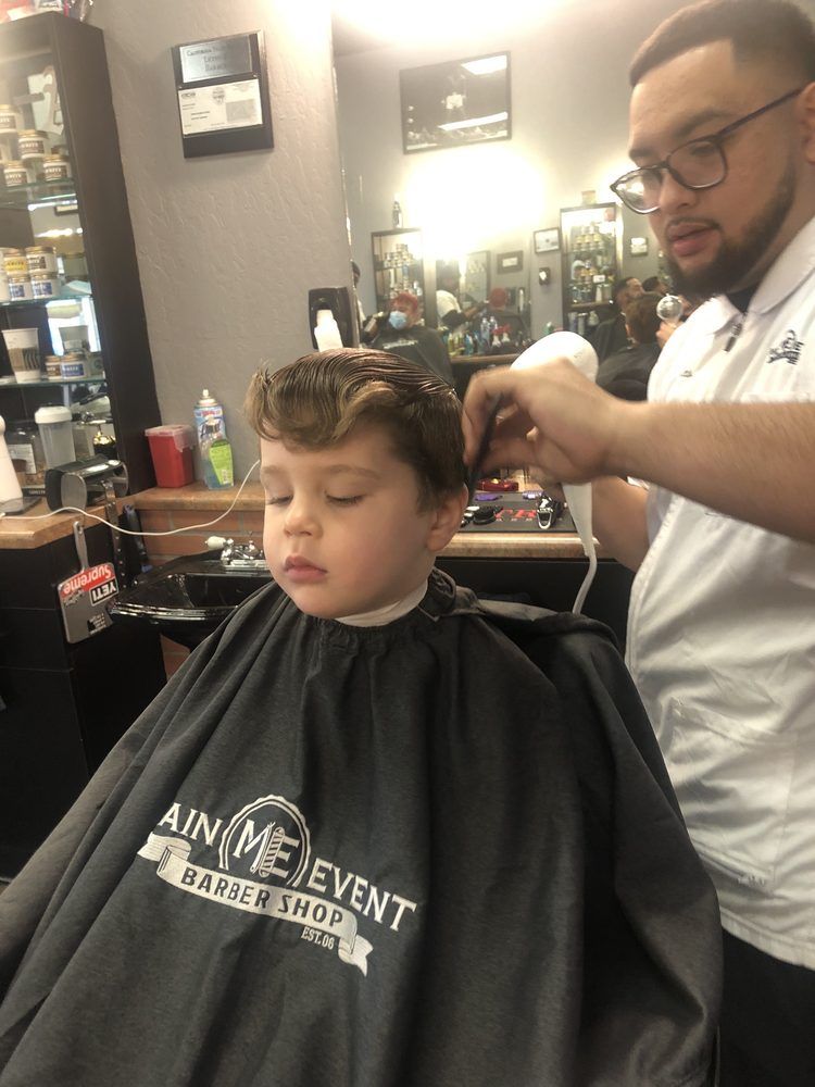A young boy with wavy hair in a Main Event Barber Shop cape while a barber styles his hair