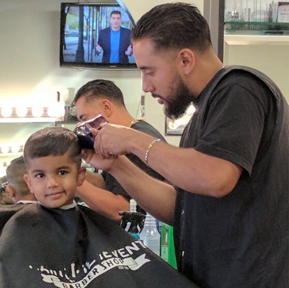 Barber Rolando Zamora clipping the side of a young boy’s hair; the boy wears a black Main Event Barber Shop cape and smiles at the camera