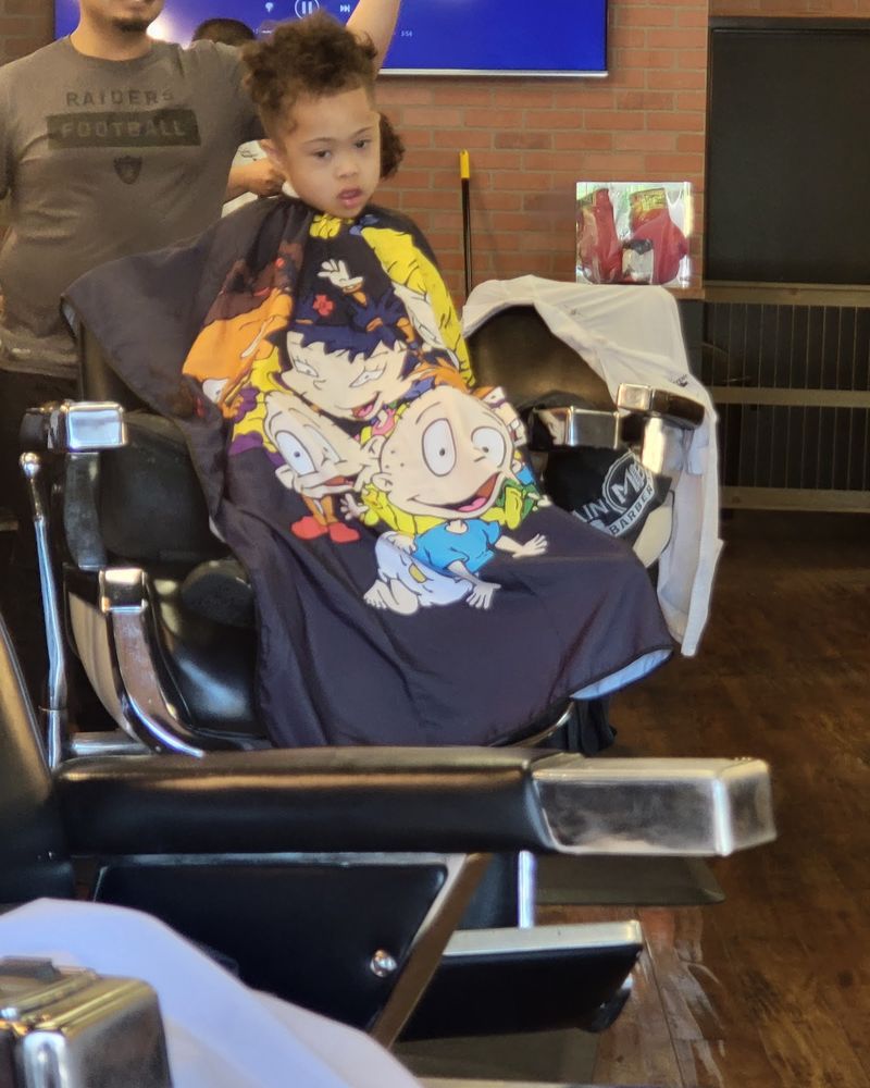 A curly-haired toddler in a barber chair wearing a colorful Rugrats cartoon cape