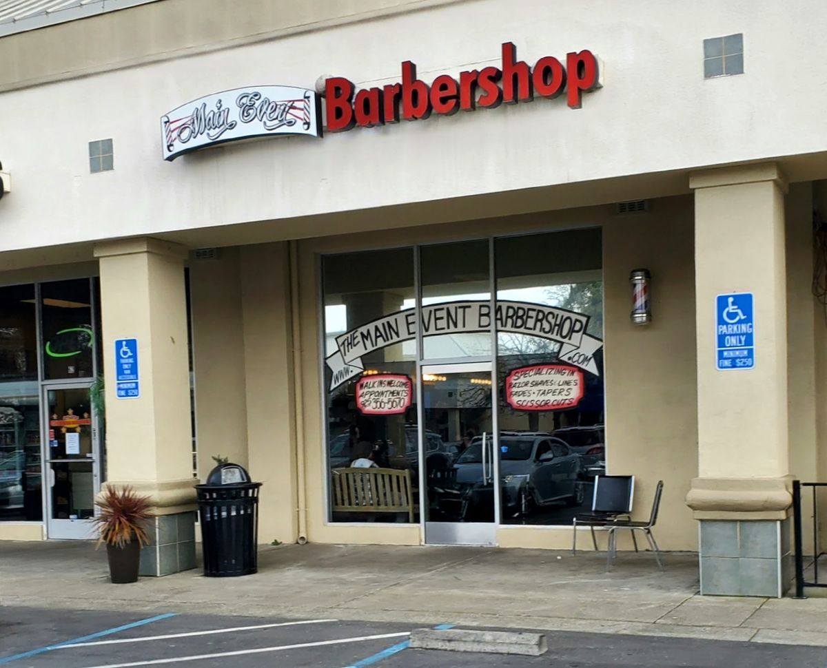 The shop front in a Concord shopping center: a red Barbershop sign above glass doors painted The Main Event Barbershop, with a barber pole, a bench and accessible parking out front